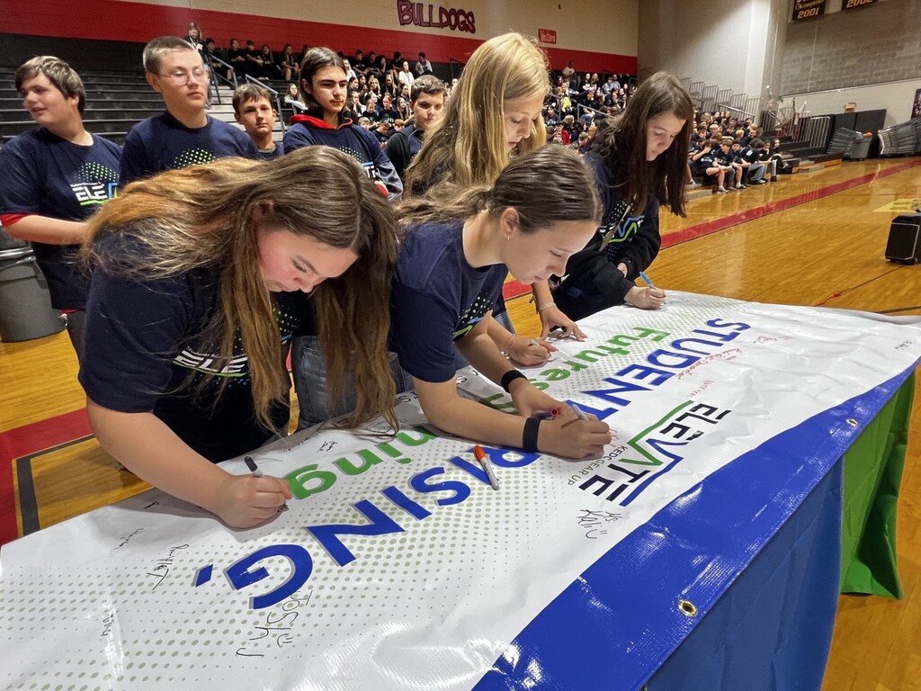 Students signing a large banner on a table at a school event, with a basketball court in the background.