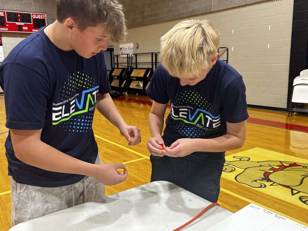 Two young people in matching blue shirts bend over a table in a gymnasium. The floor has a large yellow cartoon character.