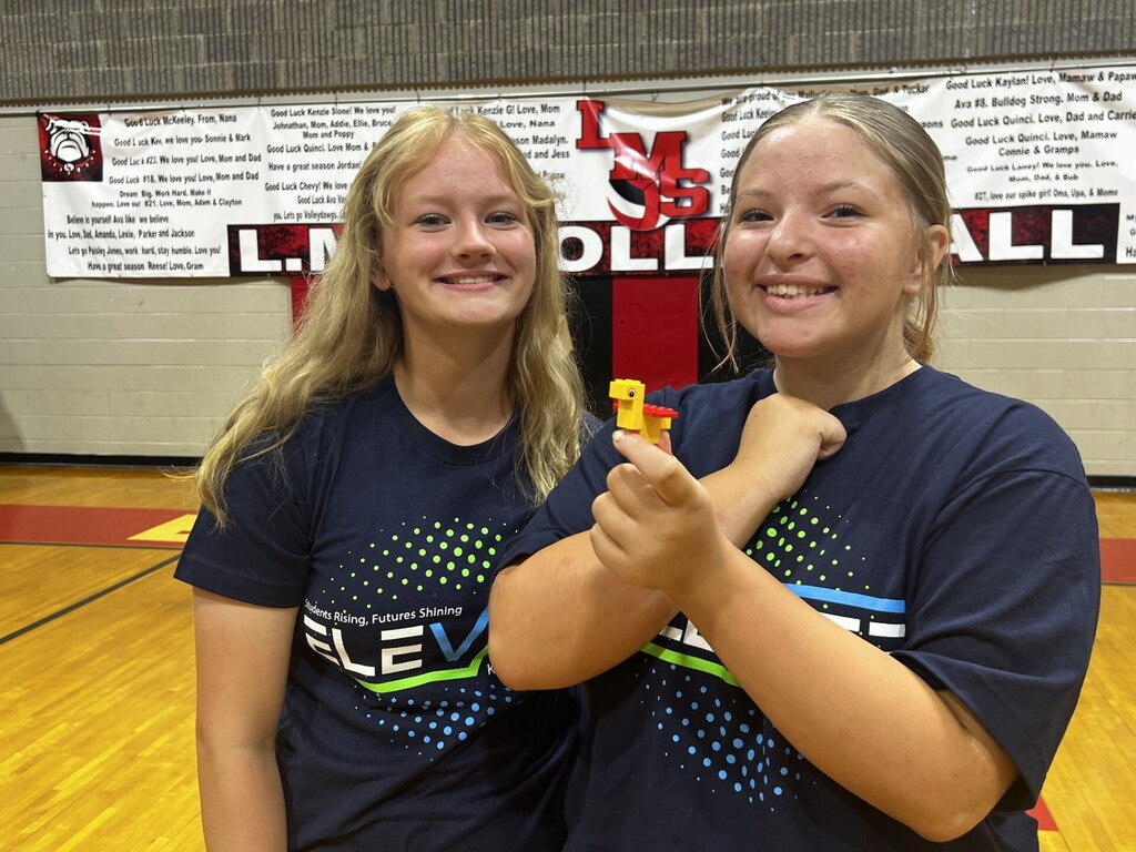 Two girls wearing navy shirts with "ELEMENT" in green letters hold up a yellow object in a gym.