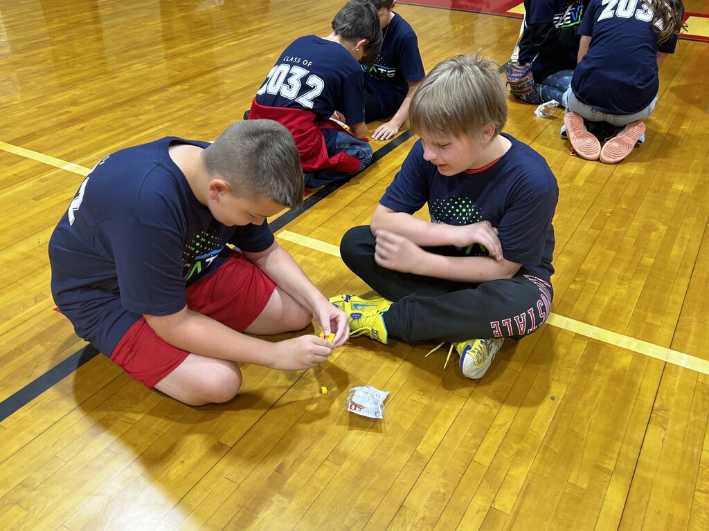 Two boys work together on a small project on a wooden gym floor. One wears a red shirt, and the other wears a blue shirt.
