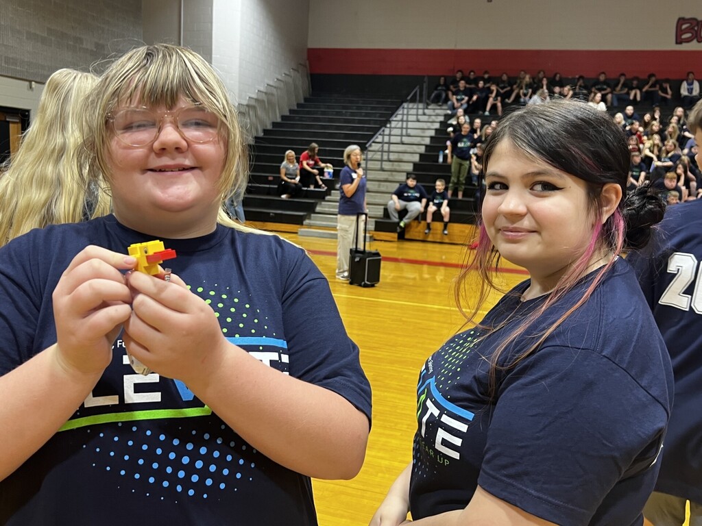 Two women with colorful hair in blue t-shirts are standing in an indoor gym with bleacher seats.