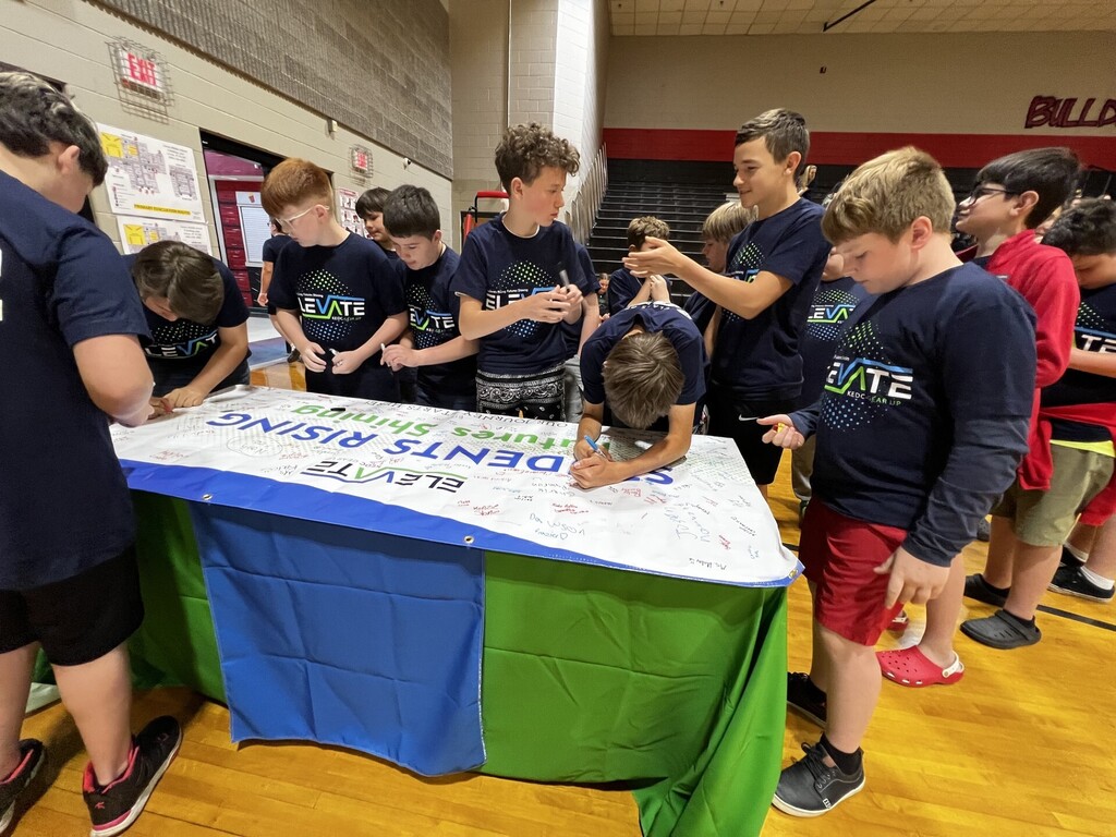 Group of boys in blue shirts gathered around a table with a white banner that reads "Join Our Clean-Up".