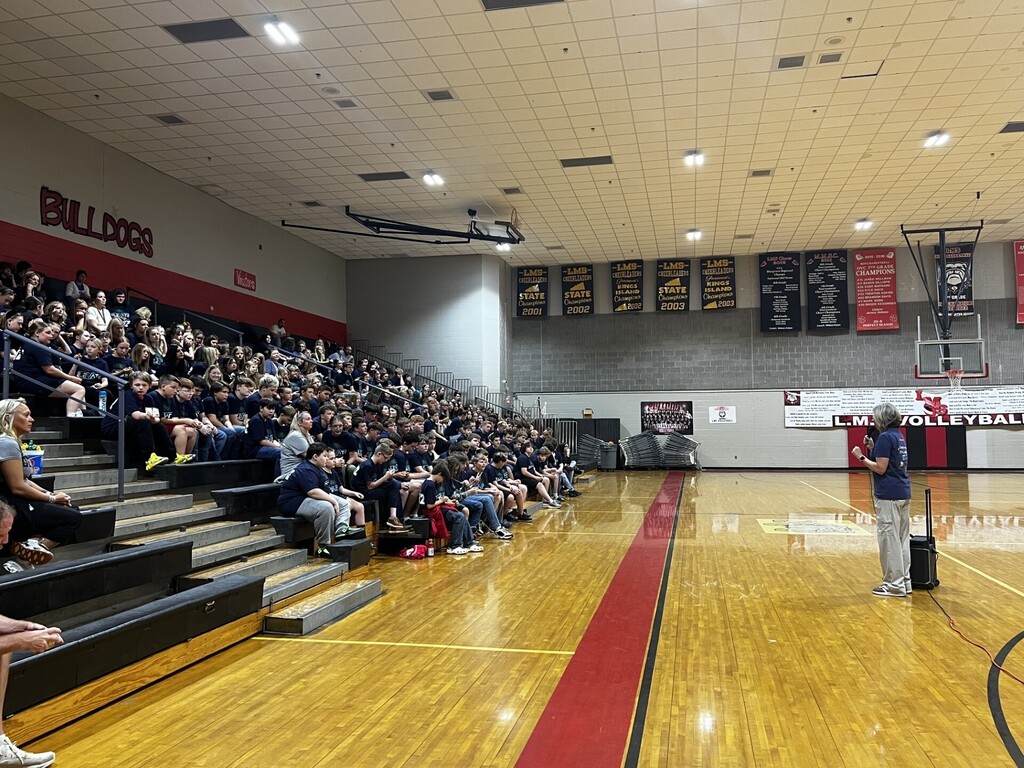 Gymnasium with wooden floors, bleachers, and spectators. A person stands near the middle with a microphone.