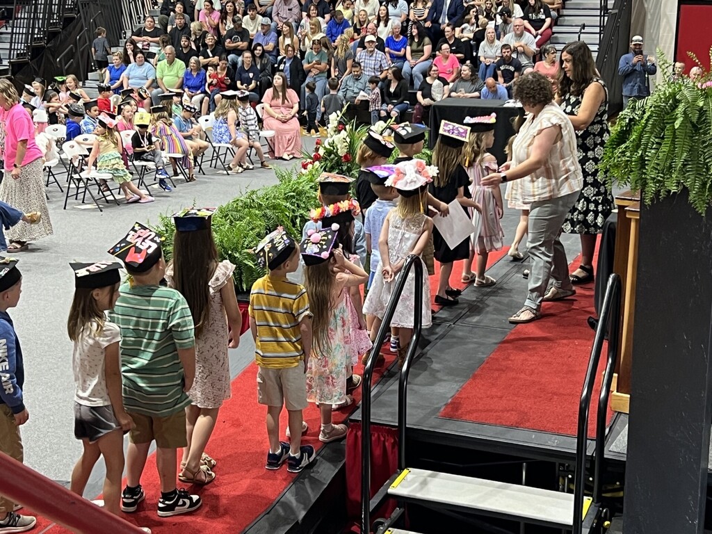 Children in graduation hats and gowns stand on stage, some holding hands, in front of an audience seated in rows.