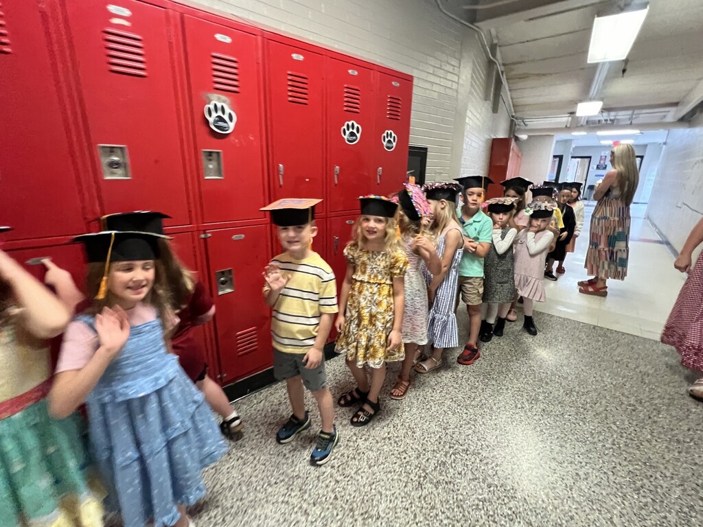 Children in caps and gowns stand in a row in front of red lockers. One person stands nearby.
