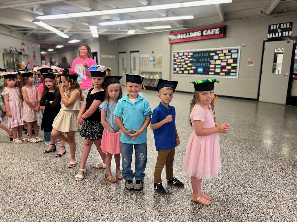 Children in caps line up in a room with a bulletin board and fluorescent lights.