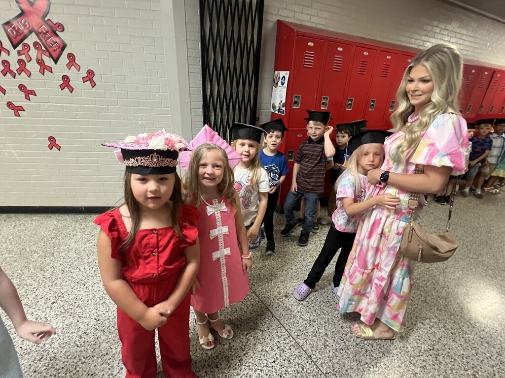 A woman stands in a school hallway with young girls, all wearing graduation caps. Behind them, red lockers line the wall.