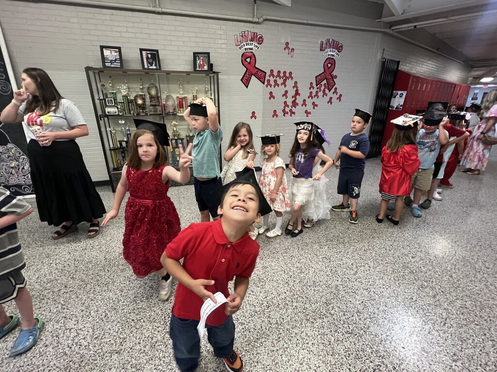 A line of children in hats and dresses walk through a room. A red ribbon decorates a white wall.