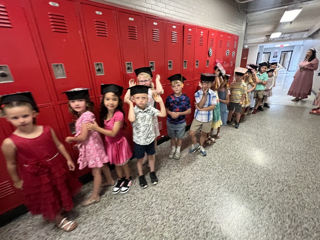 Many children in graduation hats stand in line by lockers. A woman stands in the hallway.