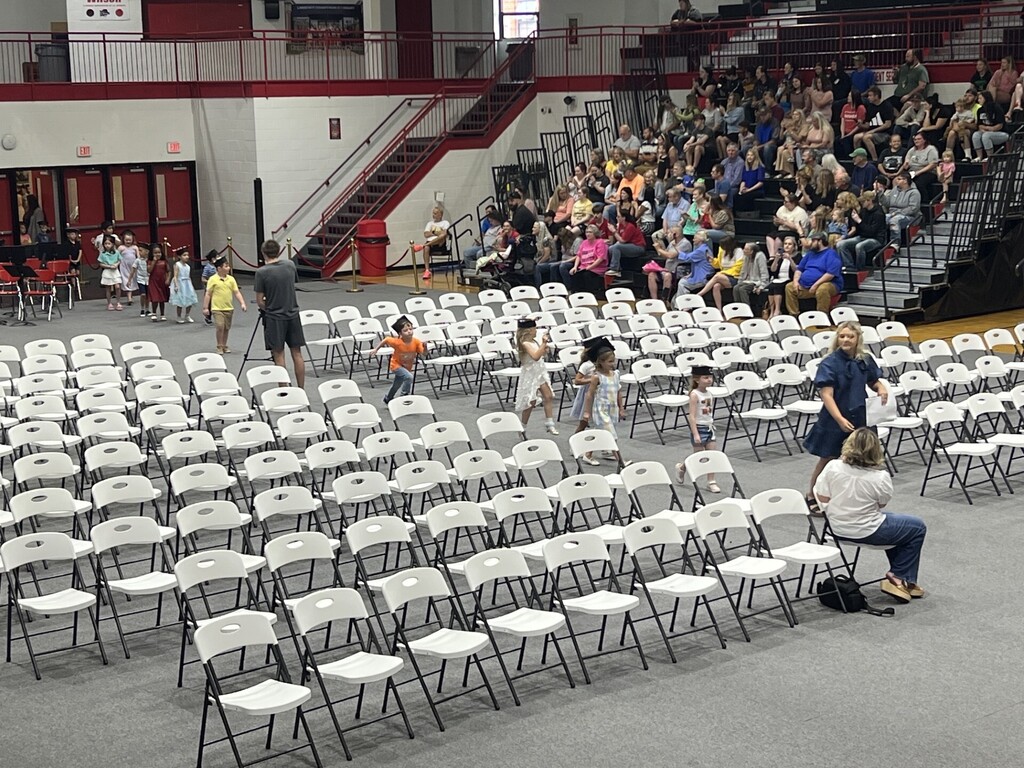 Many people are seated in rows of chairs in a large, indoor space. Some people stand and walk among the chairs.