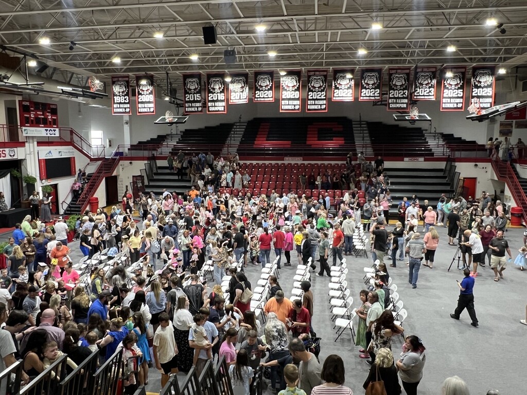 Indoor auditorium with large crowd seated in rows and standing, with banners hanging above and lights on ceiling.