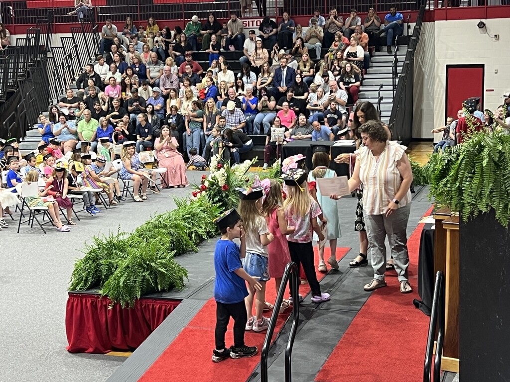 A group of children walks on a red carpet with a woman helping them. They are in front of a seated audience.