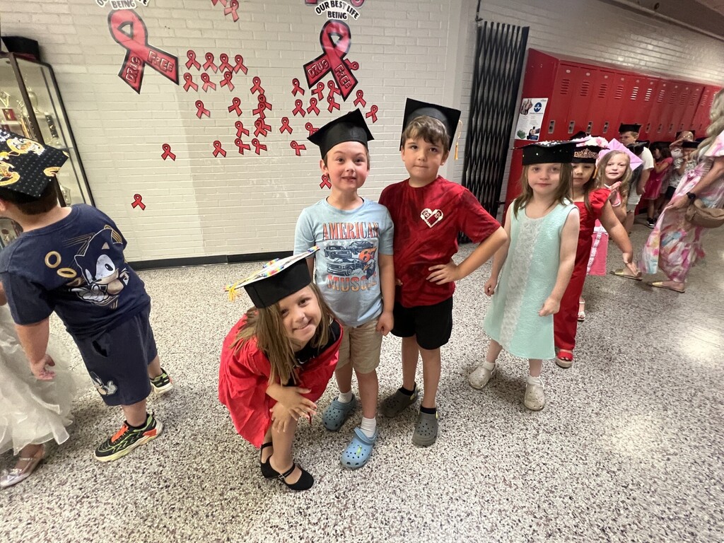 Children in caps and gowns smile in a hallway with pink ribbons on the wall.