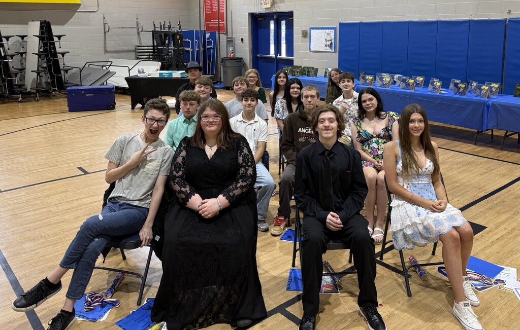A group of students and adults in a gym, some seated on chairs and others standing.