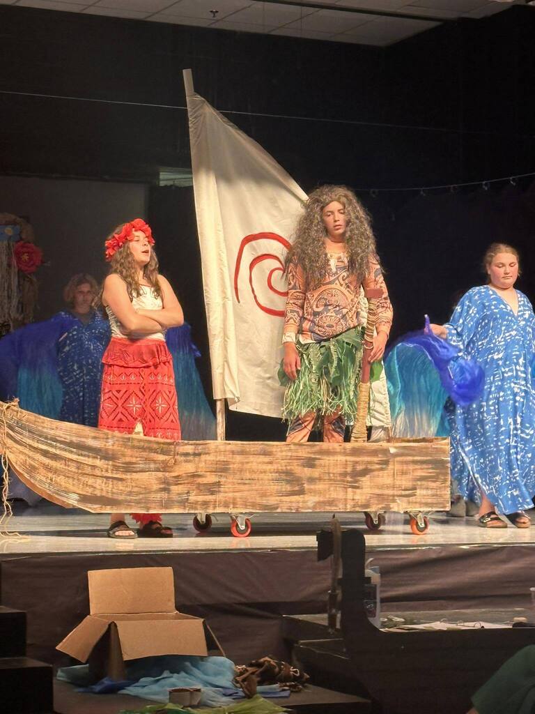 Performers stand on a stage with a boat prop. One in red, one in blue, and one in green.