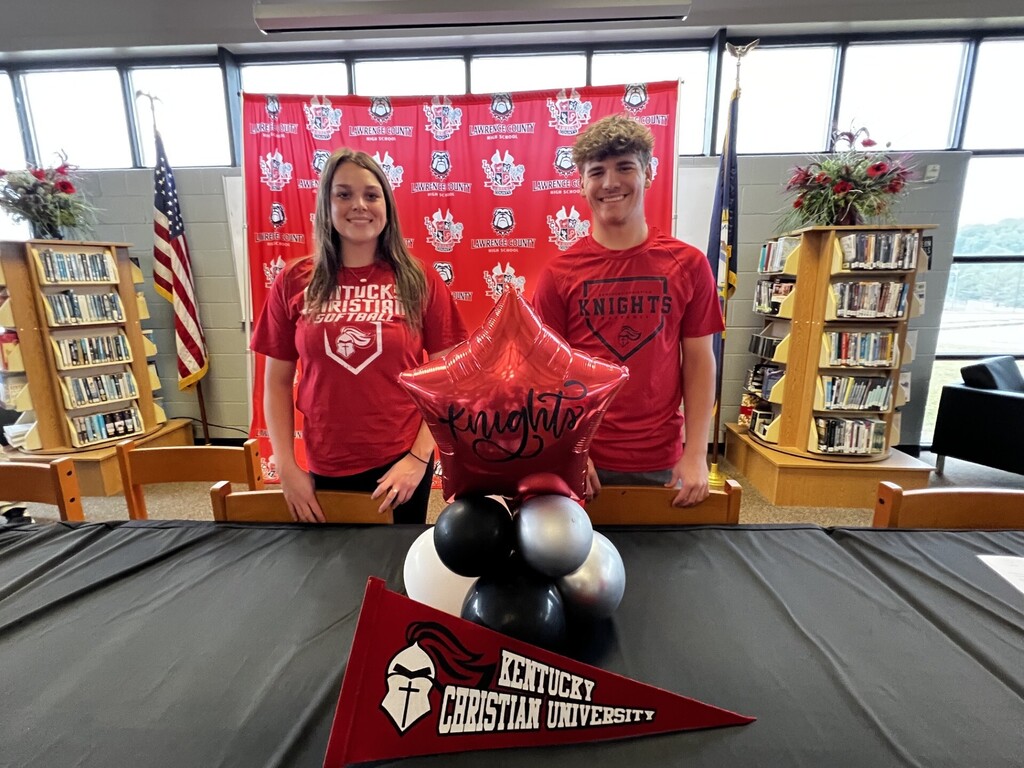 Two people in red shirts standing by a table. A red star balloon and black balloons are on the table.