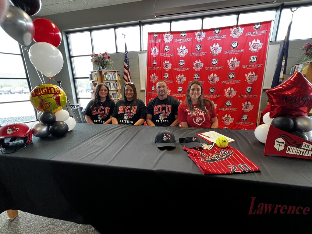 Four people in matching shirts pose for a photo at a table, with balloons and a red backdrop.
