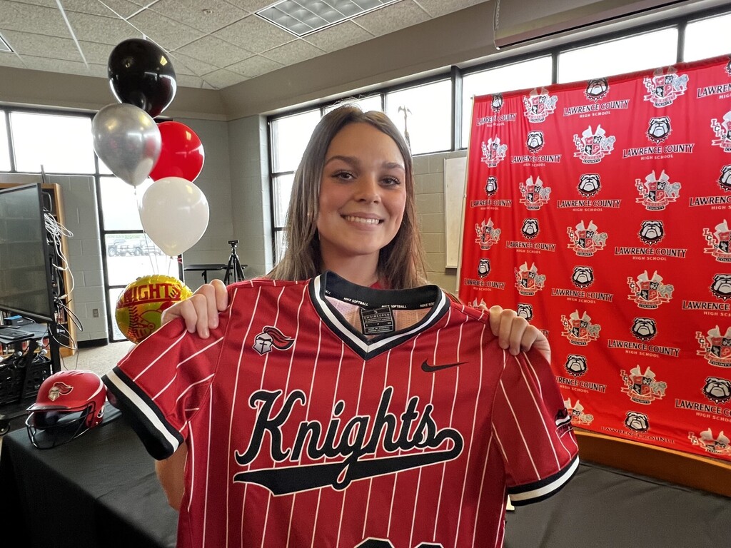 A woman smiles and holds up a red Knights jersey. Behind her is a red backdrop with black text and a table with balloons.