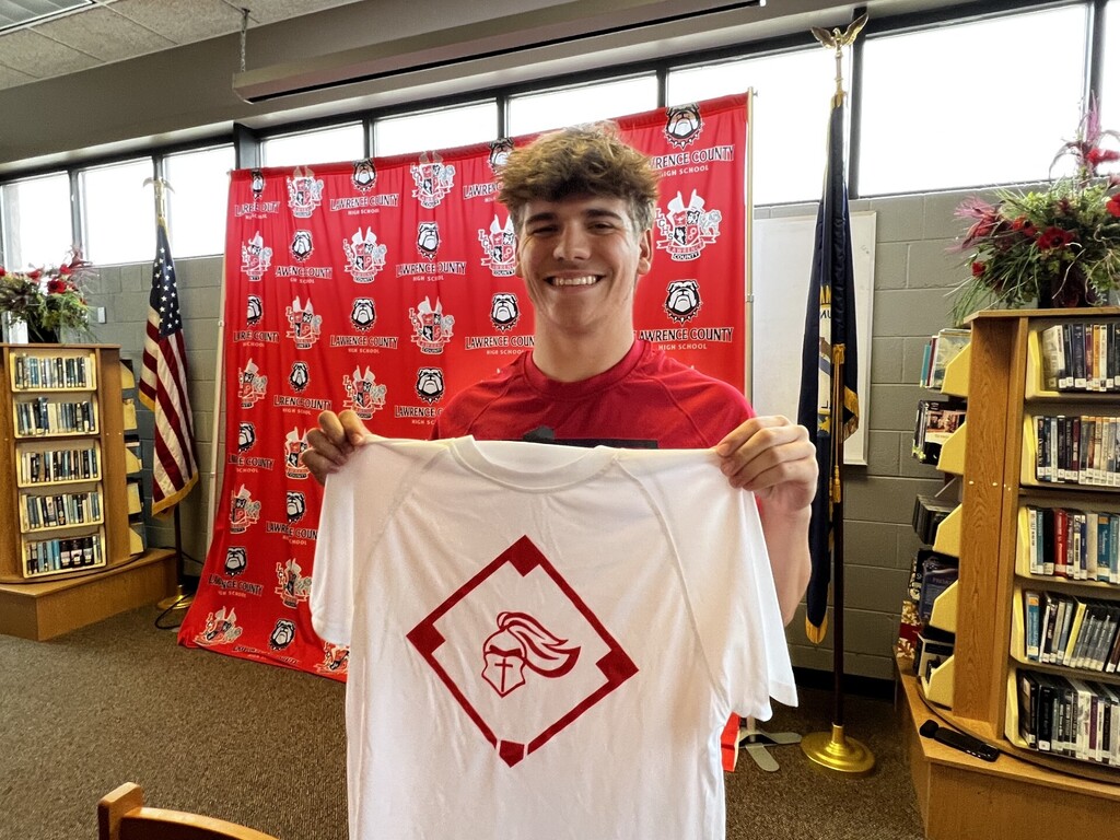 A person in a red shirt holds up a white t-shirt with a red logo in a library.