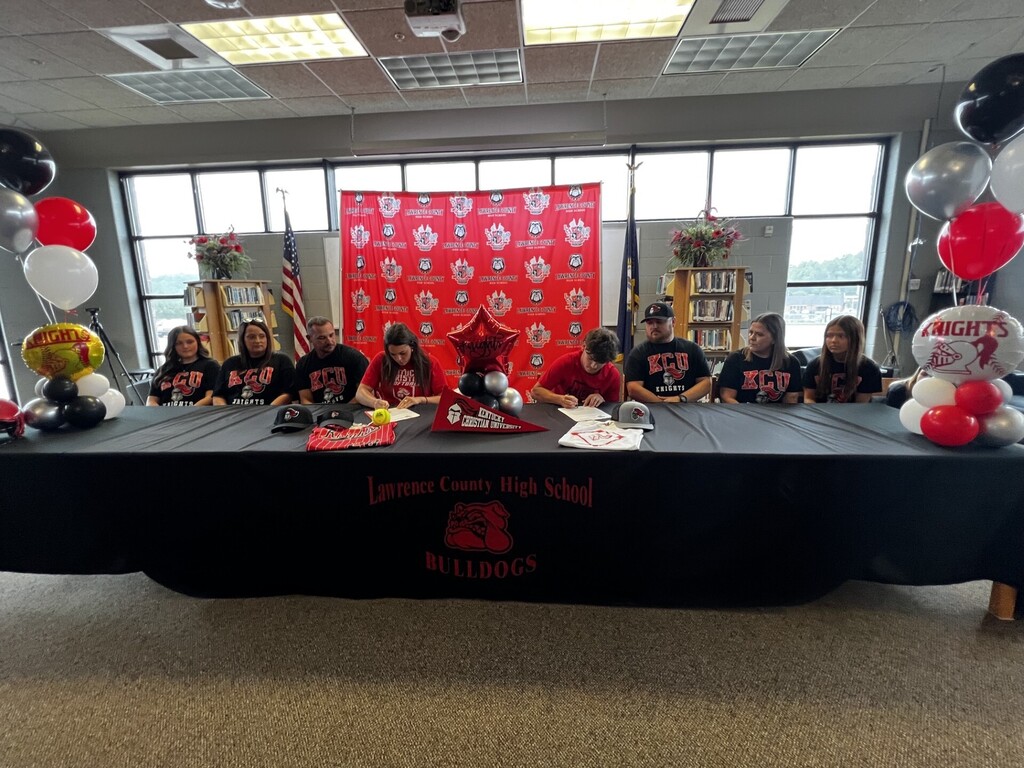 A group of people seated at a long table with a red backdrop. A banner reads "High School."