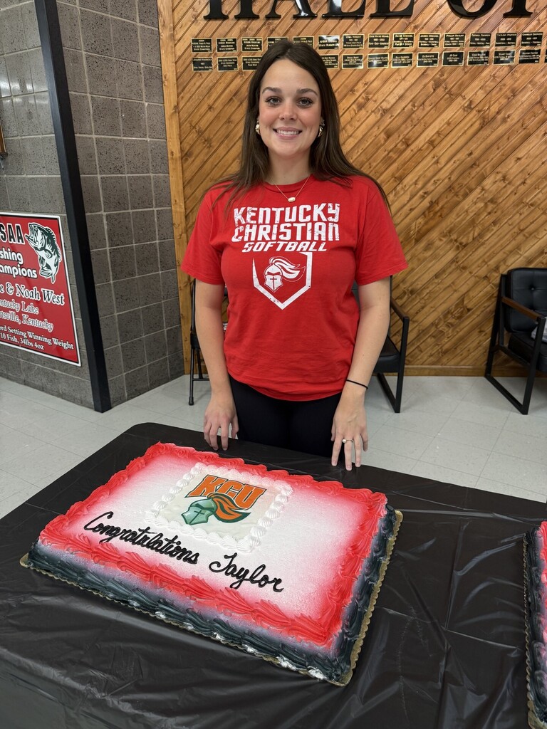 A woman in a red shirt stands beside a cake decorated with a team logo and a message.