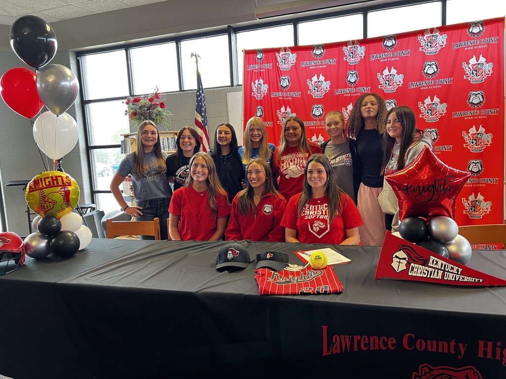A group of women in red shirts stands behind a table at a signing event. They are surrounded by balloons and a red backdrop.