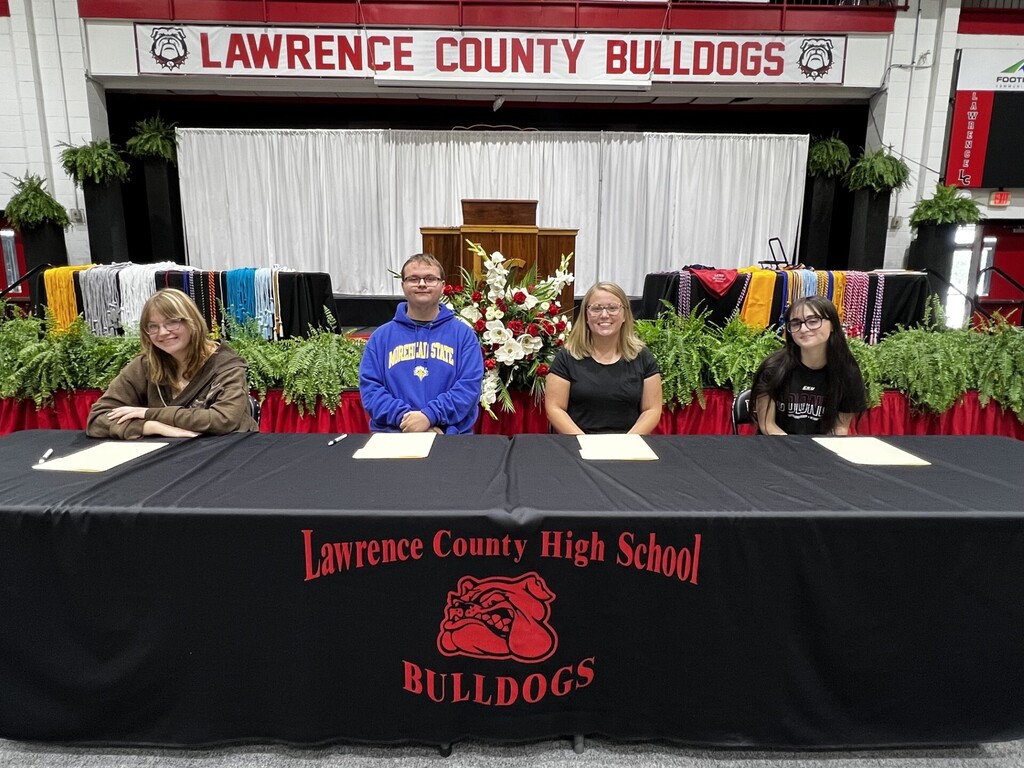Four people sit at a table with a black cover and "Lawrence County High School" logo. A stage is behind them.