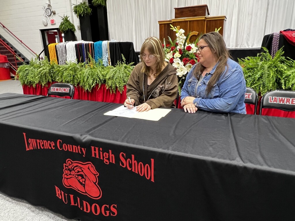 Two women sit at a table, one signing a document while the other looks on. The table has a Lawrence County High School logo.