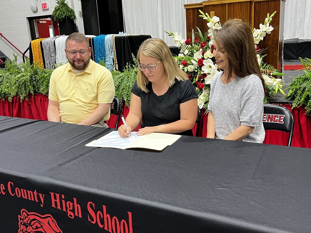 Three people, two women and a man, seated at a table with a sign that reads "County High School".