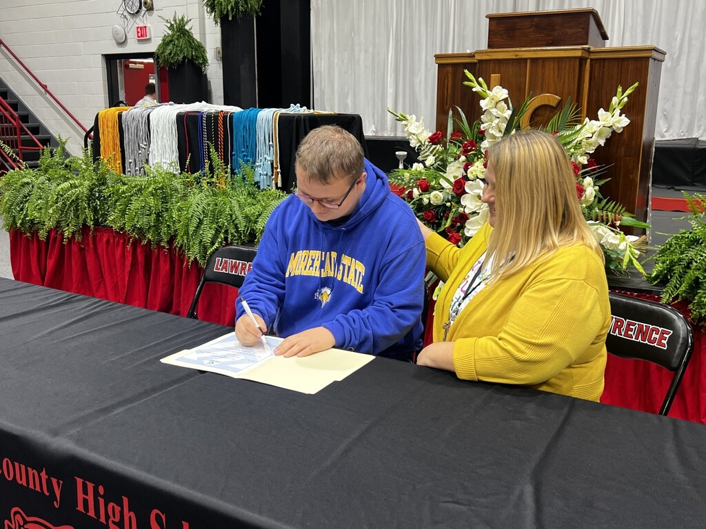 Two people seated, one signing a document on a table covered with a black cloth. Floral arrangement behind them.