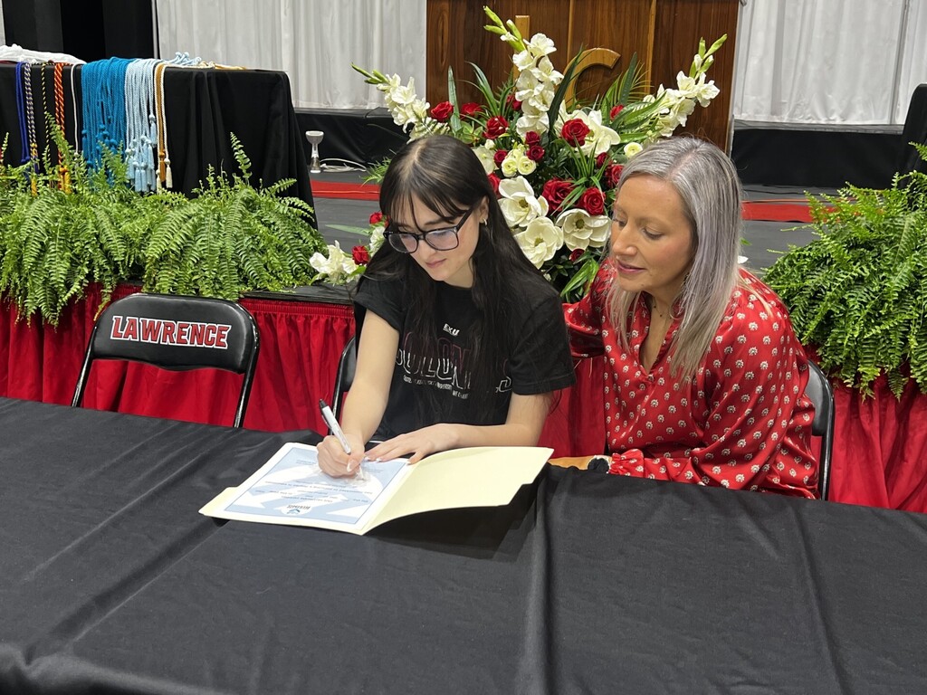 Two women at a table, one in glasses signing a book for the other woman in a red polka dot dress.