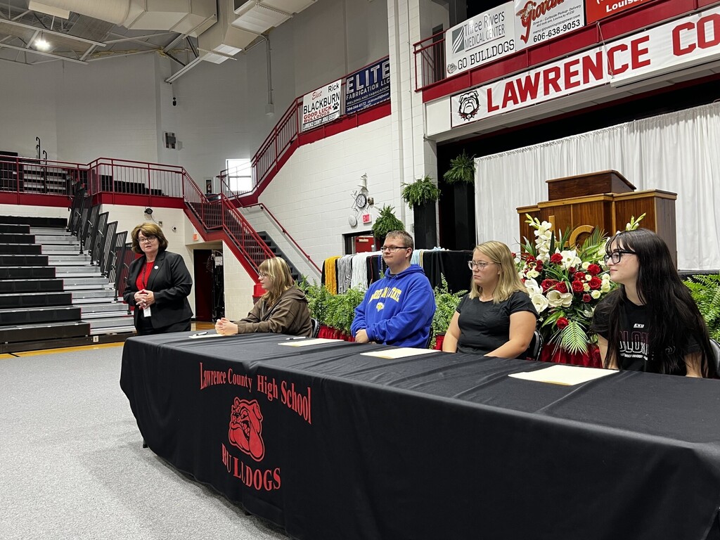 Five people are seated at a long table in a gym. A table cloth covers the table and has the words "High School" on it.