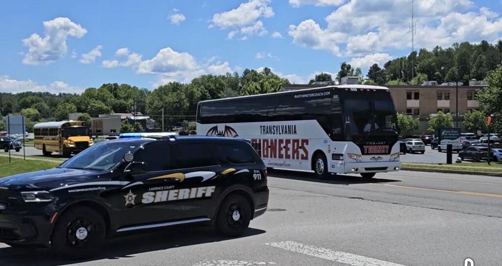 Police SUV with "SHERIFF" on side drives past a tour bus labeled "Engineers" on a street with trees and sky.