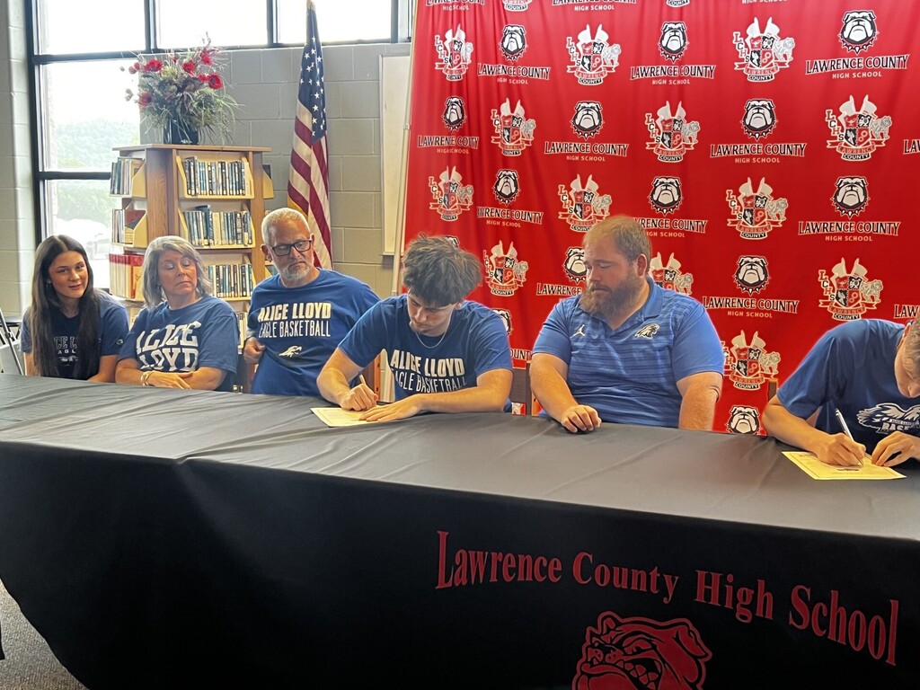 Five people are seated around a table, signing papers. Behind them, a red backdrop displays Lawrence County High School.