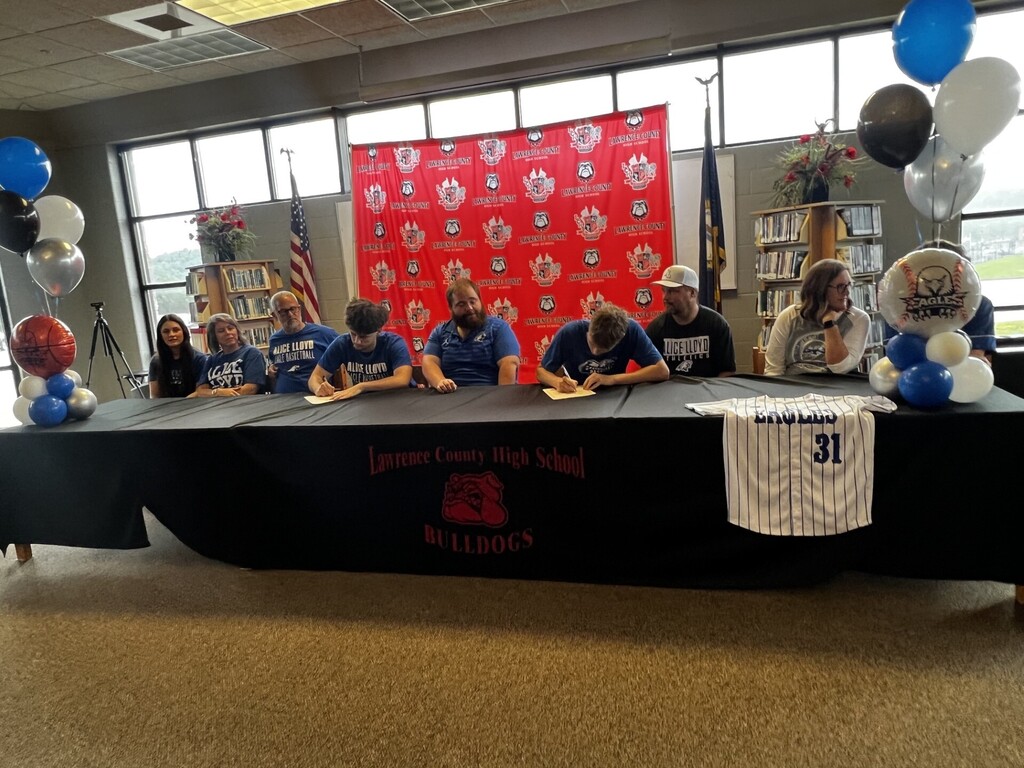 A group of people seated at a table, signing documents, with balloons and banners in the background.