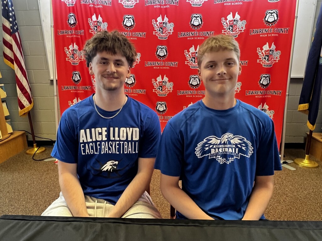 Two boys in blue Alice Lloyd University Eagles basketball shirts seated at a table with a red banner behind them.