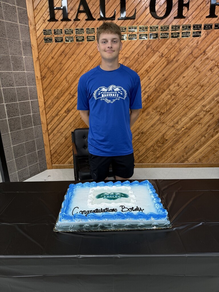 Person standing behind a table with a cake reading "Congratulations Boday" in front of a wooden wall.