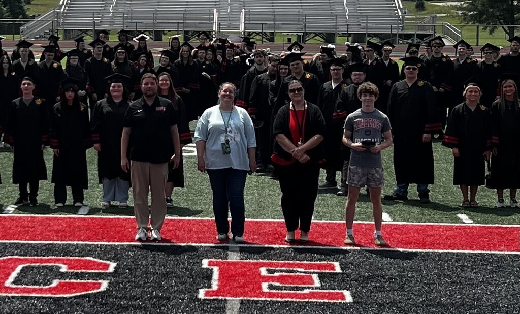 A group stands in black graduation attire on a field, with some individuals wearing sunglasses.