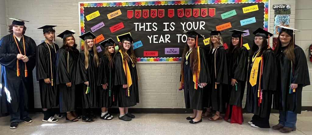 A group of people in graduation gowns, likely students, stand in front of a banner.