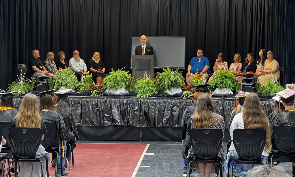 Graduates sit in chairs at a ceremony. A man speaks at a podium with a microphone. Plants decorate the stage.