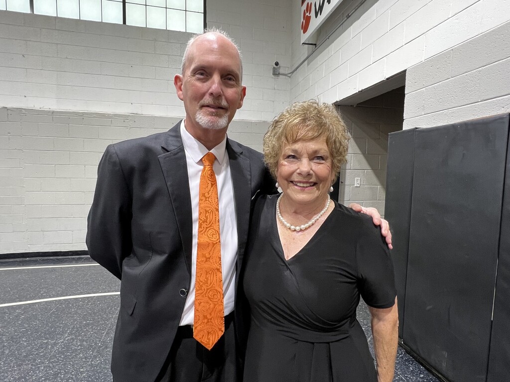 An adult man in an orange tie and a woman in a black dress stand together, smiling, in a gym.