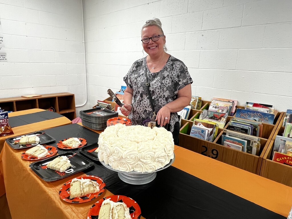 Woman stands behind a table, showing a cake with white frosting. Multiple plates hold cake pieces.