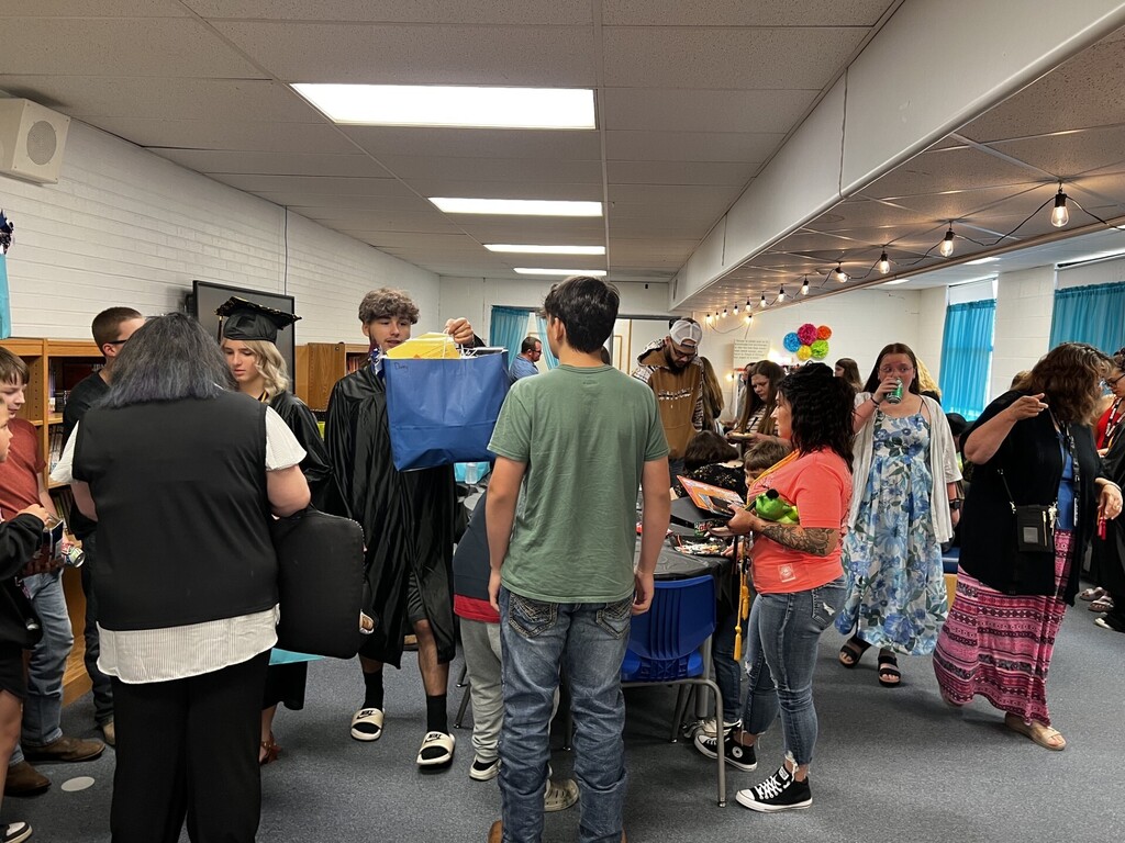 Many people in a room with tables, some wearing graduation attire and others holding items.