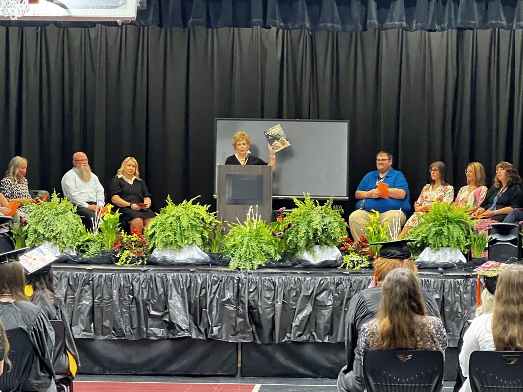 A ceremony on stage with plants; a woman speaks at a podium, and people sit in chairs.