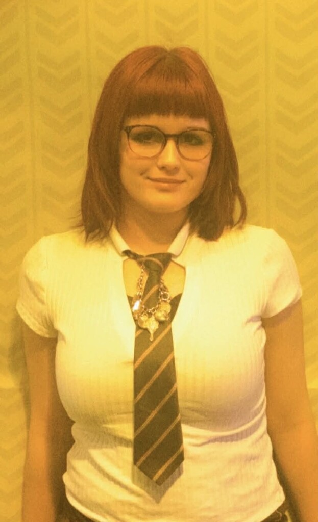 A woman with short red hair stands in front of a patterned wall. She wears a white top, a striped tie, and glasses.