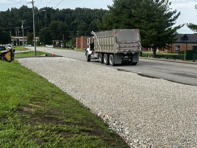 Gravel road under construction. A truck is on the road. Green grass and trees are on the side.