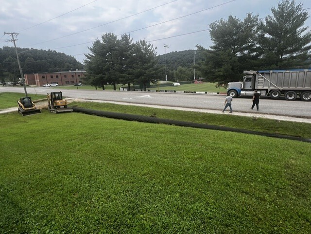 Road construction in progress. Two people and a machinery vehicle are on the road. Two machines are on the grassy lawn.