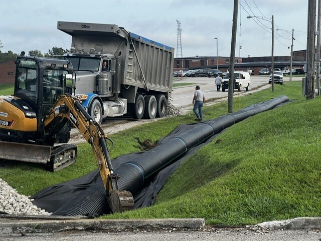 Road workers use a yellow excavator and dump truck to lay pipe. A person walks nearby, and a car is parked in a lot.