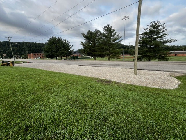 An empty street with gravel median and lush grass on either side. Trees line the road.