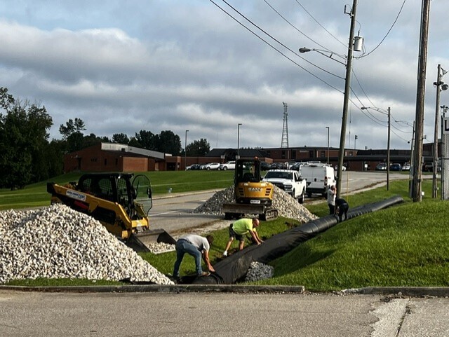 Workers lay new pavement in a grassy area by a road, with a construction vehicle parked nearby.